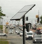 Image of Flock Camera. This camera is mounted on a black pole with a solar panel that powers the camera. 
